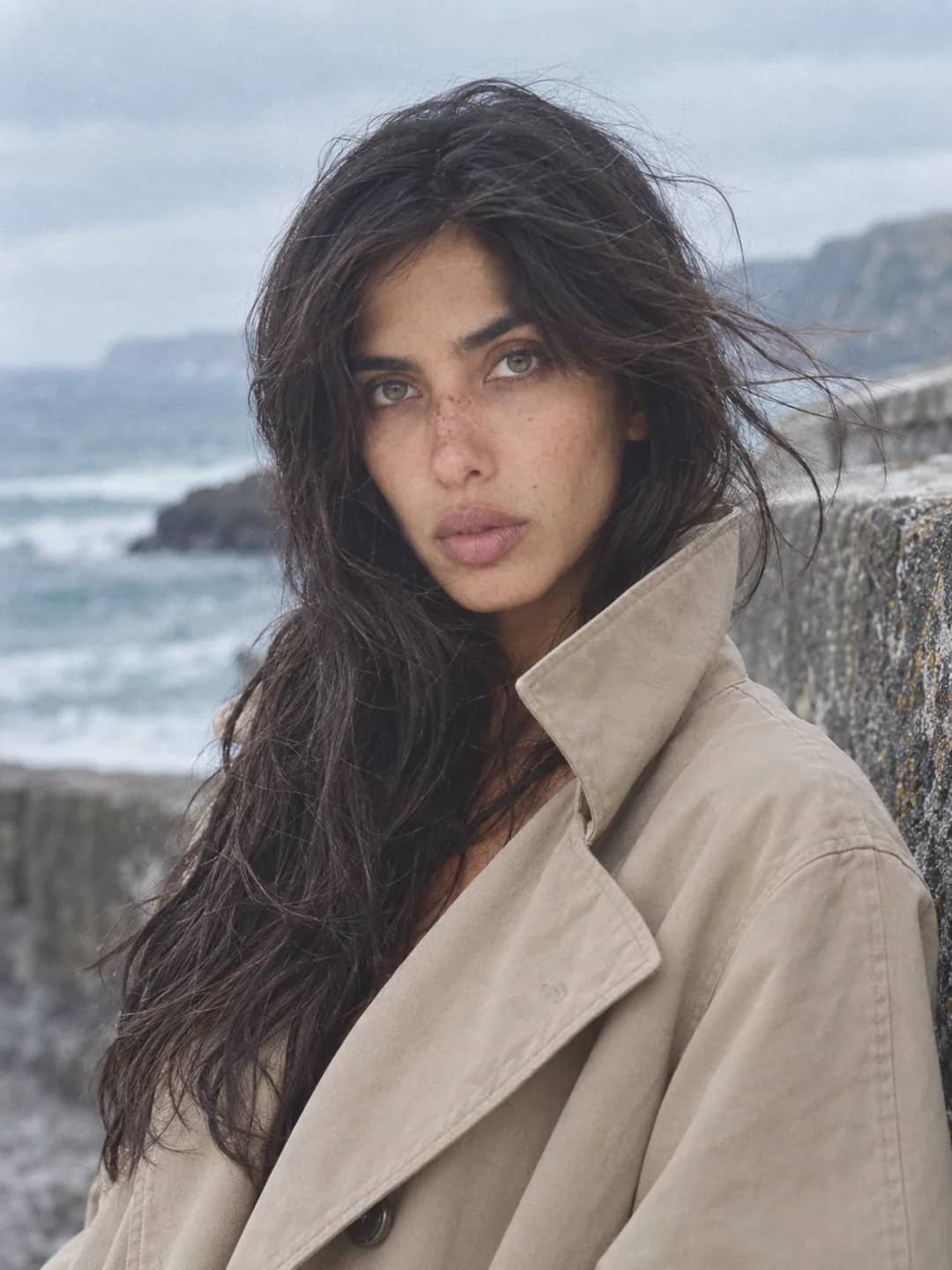 AI-generated photograph of Danielle in a trench coat on a sea wall, hair blown across her face