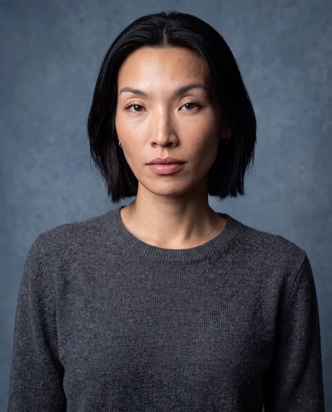 AI-generated First Look frame of Promptography cofounder Gift: a frontal studio headshot, neutral expression, charcoal crew-neck sweater against a blue-grey backdrop